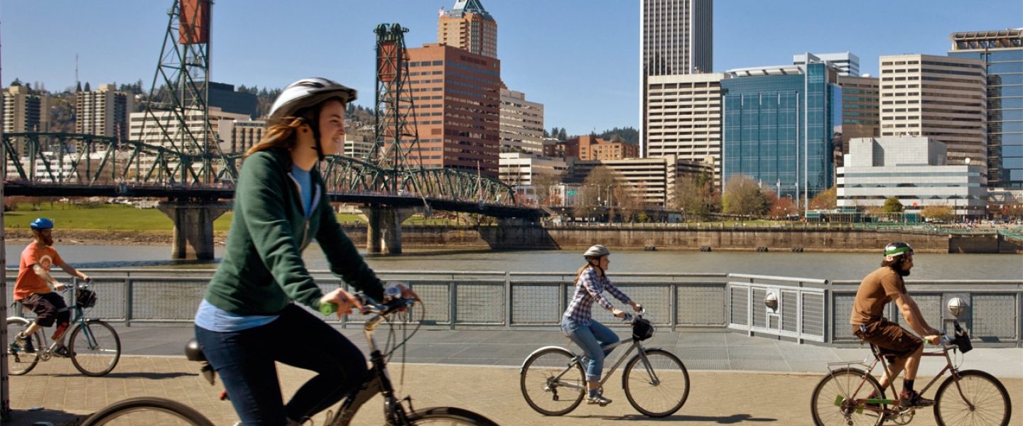 Cyclists bike along the Vera Katz Eastbank Esplanade in Portland, OR. The city skyline and Hawthorne bridge are seen in the background